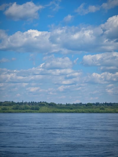 The image depicts a wide, calm river flowing through a rural landscape under a partly cloudy sky. The river occupies the lower half of the frame, its surface textured with gentle ripples reflecting the sky's blue and white. The far bank of the river is lined with a dense band of green vegetation, including trees and bushes, which forms a natural horizon. Above the treeline, the sky is a vibrant blue, adorned with scattered white cumulus clouds that create a dynamic and airy atmosphere. The scene appears serene and untouched, suggesting a tranquil natural setting. The location is identified as Vysokogorskii, Russia. There are no people or specific man-made structures visible in the image, emphasizing the natural beauty of the environment.