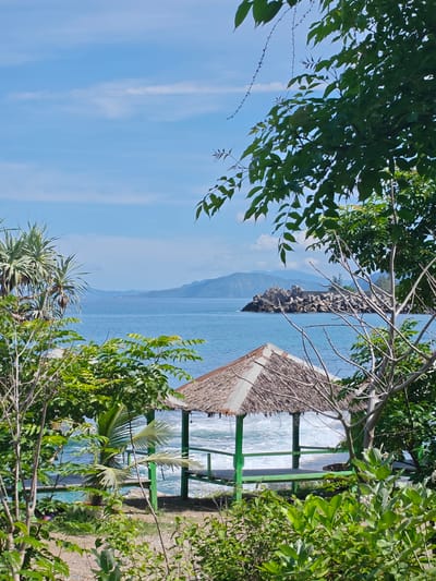 The image shows a scene from the city of Naga Umbang, Indonesia, featuring a thatched-roof gazebo located near the coast. The gazebo is painted in green, offering a vantage point overlooking the ocean. The waves are crashing against the base of the gazebo, suggesting it's located close to the shoreline. The water is blue, with an island and mountainous terrain visible in the distance. The foreground features a variety of green foliage and trees, framing the gazebo and ocean view.