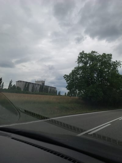 The image depicts a scene captured from a vehicle, likely from a dashboard camera, as evidenced by the reflection of the interior on the right side of the frame. The perspective is tilted, suggesting the vehicle is moving. The main subjects are a large, mature tree with dense green foliage and a long, multi-story building with a gray facade and numerous windows. The building appears industrial or agricultural, possibly a grain silo or warehouse, with antennas on its roof.
The setting is rural, with a paved road featuring white lane markings visible in the foreground on the right. To the left of the road, a grassy field and the aforementioned building are situated. Beyond the building, a line of trees stretches into the distance. The sky is overcast with heavy, gray clouds, indicating a cloudy or possibly stormy weather condition. The time of day is difficult to ascertain precisely due to the cloud cover, but it appears to be daytime.
The overall mood is somewhat somber and subdued, likely due to the overcast sky. No people are visible in the image. No text is discernible. The location context provided, Záhoří, Czechia, adds specific geographic information to the scene. The scene captures a snapshot of a journey through a rural landscape under a gray sky, with a prominent natural element (tree) and an industrial or utilitarian structure.
