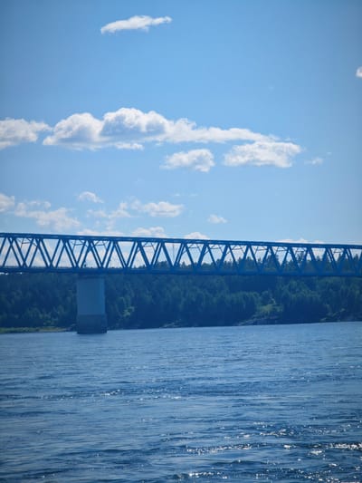 This is a photograph of a blue truss bridge crossing a body of water, with a forest-covered hillside on the far bank. The sky is blue with scattered white clouds. The water in the foreground is rippling, reflecting sunlight. The bridge appears to be a railway bridge or a road bridge of significant length. One concrete pier is visible supporting the bridge. The scene is likely located in the vicinity of Abalakovo, Russia, given the context. There are no people visible in the image, and the focus is on the bridge and the natural landscape.