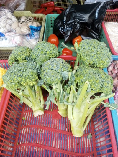 This image depicts a market stall or produce display in Naung Bo Oe, Myanmar, primarily featuring fresh produce and packaged goods within various plastic baskets. In the immediate foreground and center, multiple heads of green broccoli with their light green stems are arranged within a red plastic basket. The basket's bottom is lined with a blue and white checkered fabric. To the far left, partially visible, is yellow corn. In the mid-ground, a beige-colored plastic basket contains several clear plastic bags of spherical, light-colored packaged food items, possibly meatballs, with visible text including "KONG" and "CARA MEMASAK." Adjacent to this, red chili peppers are visible in another beige basket. Further right, a green plastic basket holds several red tomatoes and a crumpled black plastic bag. To the far right, a pink-rimmed basket contains what appears to be white, shredded food items in a clear bag, and another basket displays numerous small, reddish-brown items, likely shallots or small onions. No individuals are visible in the scene.