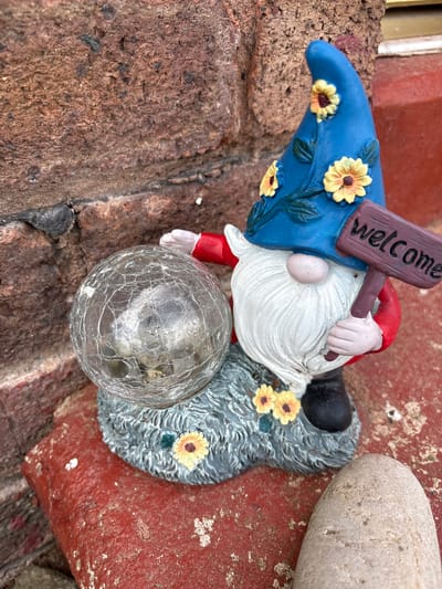 A close-up shot shows a garden gnome figurine on a rough, reddish-brown textured surface, likely brick or stone. The gnome has a blue hat decorated with yellow sunflowers, a white beard, and a red shirt. He holds a sign that reads "welcome". In front of the gnome is a cracked glass sphere. Beside the gnome is a smooth, light-colored stone. The setting suggests an outdoor location, possibly a garden or patio, and is likely located in Pulverbatch, United Kingdom.