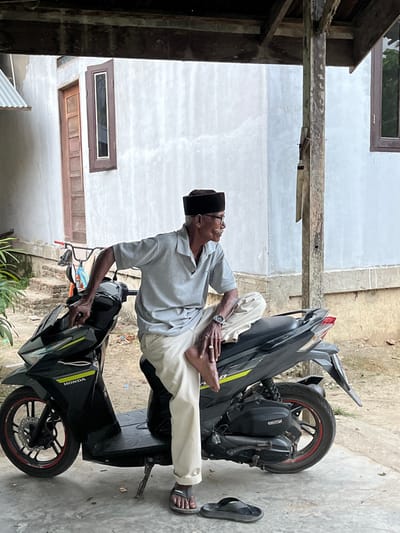 An elderly man is seated sideways on a black Honda automatic scooter, parked on a light grey concrete surface in Payakambuk, Indonesia. He wears a light grey polo shirt, light beige trousers with the right leg rolled up, a black peci, and spectacles. A wristwatch is visible on his left wrist and a ring on his right ring finger. His left foot is bare and tucked onto the scooter seat; his right foot is on the ground, wearing one dark sandal. A second dark sandal lies on the ground nearby. He looks towards the right, with his right hand resting on the scooter's handlebar area. The scooter has red accents on its wheels and a green/yellow stripe. Behind him is a light-colored, possibly white or light grey, plastered building with a wooden door and two vertical wooden-framed windows. A partially visible red bicycle leans against the building to the left of the door. Overhead, a dark wooden beam and a rough wooden support pillar indicate a covered outdoor area. Green foliage is visible on the far left.