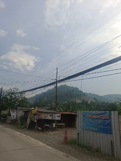 The scene is a road in Lagadar, Indonesia (latitude: -6.919, longitude: 107.538). There are a series of telephone poles with multiple power lines, a mountainside in the distance, and a road with a shoulder on the right. On the shoulder there are multiple stalls or shacks made of wood and corrugated metal. There is a blue sign with white lettering on it, advertising "BB. Bergerak Swakarsa Mandiri" on the right side of the road. The road is paved and appears to be gray in color. There is an orange traffic cone on the right shoulder of the road. The scene is overcast and there are clouds in the sky.
