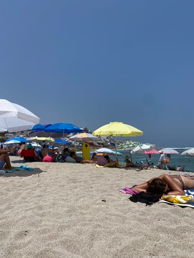 The scene takes place on a beach in Punta Hermosa, Peru, at the coordinates -12.325 latitude and -76.835 longitude. The image depicts a sunny day with clear blue skies. The beach is covered in fine, light-colored sand. Numerous people are scattered across the beach, relaxing under various brightly colored beach umbrellas. The umbrellas are arranged in a line along the shore, creating a visually appealing pattern. Many people are wearing swimsuits, and some are engaged in conversation or reading books. Several people are lying on towels, basking in the sun. A woman is lying on her stomach on a pink towel, her head resting on her arm. She is wearing a black bathing suit top and a yellow bottom. A surfboard is propped up against a sand dune in the background, indicating that this may be a popular surfing spot. The ocean is visible in the background, with a few surfers riding waves in the distance. The overall atmosphere is one of leisure and relaxation.