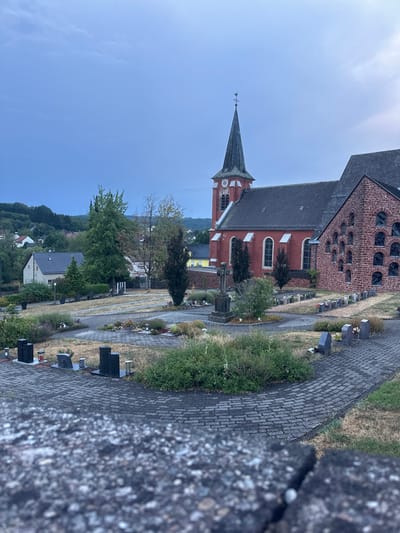 The image depicts a cemetery adjacent to a church in the city of Wadern, Germany. In the foreground, a grey stone wall is visible. Beyond the wall, paved pathways wind through the cemetery, lined with gravestones of various sizes and shapes. Some gravestones are dark, rectangular obelisks, while others are smaller, grey upright stones. There are also some grave plots with low-lying plants and flowers. A large, stone crucifix stands prominently in the middle ground.
To the right of the cemetery is a section of the church constructed with red brick, featuring a facade with numerous arched windows. Adjacent to this brick section is the main body of the church, painted red, with a tall, grey spire topped with a weather vane. The church has large, arched windows with white frames.
In the background, a residential area with houses can be seen nestled among rolling, tree-covered hills. The sky is overcast with a pale blue hue, suggesting it is either early morning or late evening. There are no people visible in the image. The overall scene is one of a quiet, rural cemetery and church setting.