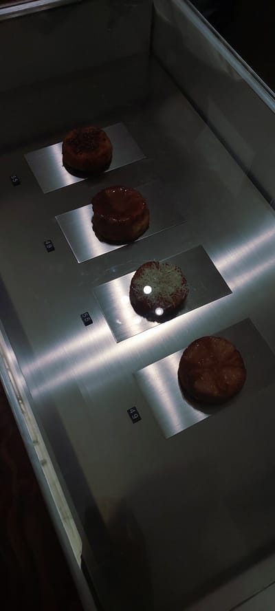 In a display case, four small, round baked goods are arranged on silver platters. The display case appears to be in a food establishment, possibly a bakery or cafe. The baked goods vary in appearance. The top one is sprinkled with dark crumbs. The second from the top is glazed with a dark, shiny topping. The third from the top is topped with shredded white material, possibly cheese or coconut. The bottom baked good has a cross-hatch pattern on its surface and a slightly glossy sheen. Small black labels with white numbers are visible below the first, second, and third items, indicating prices of 55, 49, and 55 respectively. The lighting in the case creates reflections on the silver surfaces and the baked goods. The background is dark and indistinct, suggesting the surrounding environment is dimly lit. The location is identified as Lengkong Kulon, Indonesia.