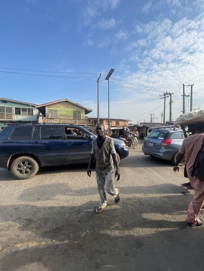 The image depicts a street scene in Akowonjo, Nigeria, under a bright, partly cloudy sky. In the foreground, a man wearing a checkered shirt and light-colored pants walks towards the viewer. He has a dark complexion and is wearing sandals. To his right, another person, partially visible and from behind, is wearing pink trousers and carrying a large bag of goods on their head, along with a backpack.

Dominating the left side of the frame is a dark blue SUV, its windows tinted. Inside, the driver and at least two passengers are visible. The road surface appears to be asphalt with some patches and debris.

In the background, several buildings are visible, including a light blue structure with balconies and a greenish-yellow building with a sloped roof. Electrical poles with numerous wires are a prominent feature of the skyward view, along with a modern street lamp. Further in the background, there are more buildings, market stalls, and a motorcycle with a rider. The overall impression is of a busy, sunlit urban environment with pedestrian and vehicular traffic.