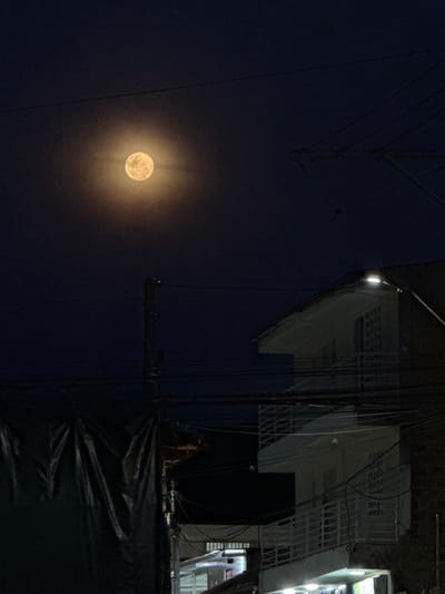 This nocturnal image depicts a street scene in Timaná, Colombia. A large, luminous full moon, surrounded by a prominent golden-yellow corona, is centered in the upper third of the dark, clear sky. Below the moon, a complex array of dark utility wires crisscrosses the frame horizontally and diagonally, connected to a dark vertical utility pole visible on the left side.

On the mid-right, a multi-story building with light-colored walls is present, featuring at least two balconies equipped with light-colored railings. The building's angled roofline is visible on the upper right. A bright white LED streetlight, mounted on a diagonal pole, extends from the right edge, illuminating the area in front of the building's upper section. The lower portion of the building shows areas of artificial illumination, suggesting interior lighting. In the lower-left quadrant, a dark, draped, sheet-like object obscures an underlying structure next to the utility pole. No individuals are visible, and no specific actions are discernible within the frame.