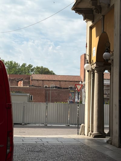 The image depicts an urban scene in Novara, Italy, likely in a historically significant area. In the foreground, a section of a red vehicle is visible on the left. The ground is paved with cobblestones and large stone slabs. A metal fence stretches across the midground, indicating a construction or renovation site behind it. Beyond the fence, red brick buildings with tiled roofs are prominent, suggesting older architecture. Several trees with dense green foliage are visible behind these buildings.
On the right side of the image, a colonnade with large stone pillars frames the view. The pillars support an arched structure with a yellow-painted facade. A traditional lamppost with a spherical white globe is mounted on the facade. Above the archway, decorative architectural elements are visible. A triangular yield sign is positioned near the lamppost, indicating a traffic regulation. In the sky, several birds are flying. The overall impression is one of an outdoor public space, possibly a street or piazza, with elements of both modern utility (the fence) and historical architecture.