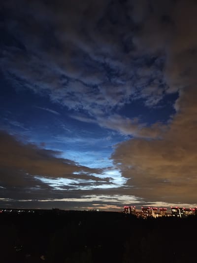 The image captures a twilight sky over the city of Vatutinki, Russia, with a line of dark trees in the foreground. The sky is a dramatic mix of dark blue and muted orange clouds, with streaks of bright blue light visible in the upper portion. In the distance, several tall buildings are illuminated, their windows glowing with warm light, and red lights are visible on their rooftops, indicating they are likely apartment complexes or office buildings. The city skyline is silhouetted against the darkening sky. The overall impression is one of a peaceful evening settling over an urban landscape.