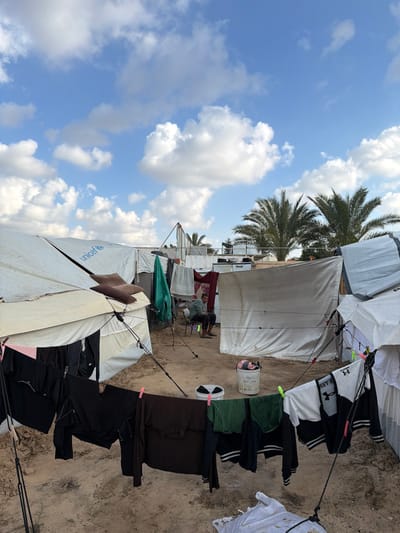 The image depicts an outdoor temporary settlement in a-Zawayda, Palestinian Territory, under a clear blue sky with scattered white cumulus clouds. The ground is composed of sand or packed earth. In the foreground, multiple clotheslines are strung across the scene, laden with various articles of clothing, including black shirts, dark trousers, green fabric, and a white hoodie with "UNDER ARMOUR" branding, all secured with clothespins. A white plastic bucket containing fabric and a small white dish with dark items are present on the ground.

Several white canvas tents serve as shelters, secured with ropes and stakes. The "unicef" logo is visible on the upper left tent. In the mid-ground, an adult male is seated barefoot on a green folding chair, facing right, and looking down at an object held in his hands. Behind him, additional tents and structures, some appearing to be made of rigid white panels, are visible. Green and maroon cloths hang from lines between these structures. In the background, several palm trees are prominent, along with further temporary structures and what appear to be containerized units topped with equipment.