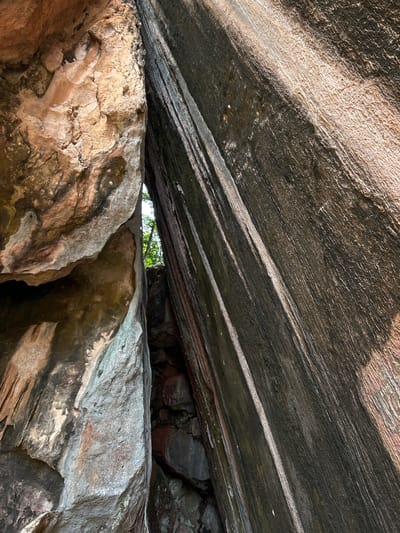The image shows a narrow crevice between two large rock formations. The rock on the left is rough and weathered, with earthy tones ranging from light brown to gray. It has a textured surface with visible cracks and indentations. The rock on the right is darker, with a smoother, more striated surface. It appears to be a darker, possibly basaltic rock, with prominent vertical striations and streaks of lighter material. Sunlight is illuminating parts of the rock faces, creating areas of light and shadow. In the background, a sliver of bright sky and some greenery, likely trees, are visible through the crevice. The foreground shows the base of the rocks, with smaller stones and debris. The scene is natural and appears to be an outdoor environment, likely a canyon or a rocky overhang. There are no people or any man-made objects visible in the image. This location is likely within the natural landscape of Krasom, Thailand, given the description of the geology and vegetation.