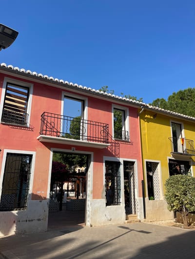The image displays a vibrant street scene featuring two adjoining, brightly colored buildings under a clear blue sky. The building on the left is painted in a warm salmon-pink, while the one on the right is a cheerful yellow. Both structures have traditional white-trimmed, red-tiled roofs and multiple windows with decorative black metal railings on balconies or in front of the windows.

On the pink building, several windows are visible. One upper-floor window features an ornate black balcony railing, casting a crisp shadow on the wall. Another lower window also has a decorative black railing. Faintly visible above one of the windows on the pink building is the number "3".

The yellow building also has several windows. From one upper window, a small yellow and red striped flag, possibly a Spanish flag, is draped over a railing. Above this window, a faint number "2" can be seen. Below it, a window is surrounded by decorative white and blue patterned tiles.

Green foliage from trees is visible along the top edges of the image and peeking over the roofs, suggesting lush surroundings. A paved street or alleyway runs along the right side of the buildings, illuminated by bright sunlight. Strong, well-defined shadows, including the elongated shadow of a streetlamp, are cast across the street and buildings, indicating midday or early afternoon. The weather is sunny and clear. No people are visible. The setting is a picturesque street in Picanya, Spain, characterized by its colorful architecture and bright light.