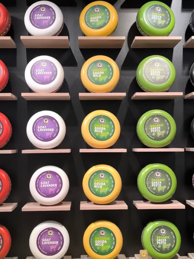 The image depicts a display of cheese wheels in a store, likely located in Leidschendam, Netherlands, based on the Dutch language labels visible on the cheese. The background is a dark, possibly black, wall.
The cheeses are arranged on small, light-colored wooden shelves that are evenly spaced. There are three distinct types of cheese displayed in vertical columns. From left to right, they are: "Goat Lavender" in white packaging, "Organic Gouda Mild" in yellow packaging, and "Organic Green Pesto" in green packaging. The cheese wheels appear to be similar in size. The packaging includes text in both English and Dutch, along with images of the animals associated with the cheeses (goats and cows). Some of the cheese wheels have price tags on the bottom right, and there's another cheese visible on the far left and right side of the image, partially cut out of the picture.