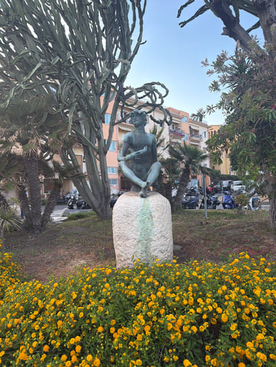 The image depicts a bronze statue of a seated male figure on top of a stone in Chianchitta, Italy. The statue is located in a park or public space with various plants, including a large cactus-like plant, palm trees, and a dense patch of yellow flowers. A building with apartments and parked motorbikes can be seen in the background.