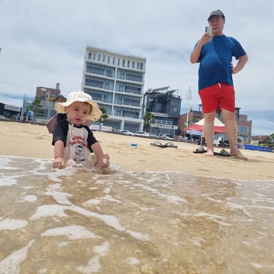 A low-angle, full shot captures a young child sitting in the shallow water at a sandy beach, with an adult man standing behind them and looking at a smartphone. The child, wearing a white sun hat with red polka dots and a black and white t-shirt with a bear graphic and the words "Aloha Sun Bear," is in the foreground, partially submerged in foamy water. Their hands are in the sand, and they appear to be playing. The adult, to the right, is wearing a blue t-shirt and red shorts and has a gray baseball cap on. He is holding a smartphone up, seemingly taking a photo or recording a video. The background features a cloudy sky, a beach, and several buildings, including a multi-story white building with many windows and a darker building with a more modern design. The scene appears to be a coastal area in Gangneung-si, South Korea, possibly near a resort or residential area. The overall atmosphere is casual and relaxed, suggesting a family outing at the beach.