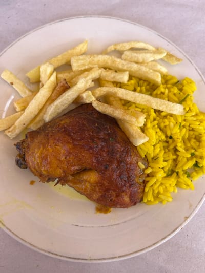 A white plate with a golden-brown chicken leg, yellow rice, and french fries. The plate is set on a textured surface. The food appears to be a meal served at a restaurant in the city of Ouarzazate, Morocco, located at the coordinates 30.924 latitude and -6.925 longitude.