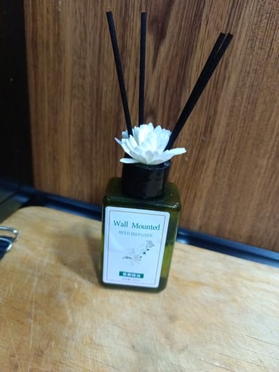 A reed diffuser with a white floral accent sits on a wooden surface, with a wooden wall in the background. The diffuser is a green glass bottle with a white label that reads "Wall Mounted REED DIFFUSER" and some Asian characters. Three black reeds are inserted into the top of the diffuser, and a white artificial flower is placed around the base of the reeds. The lighting suggests it is daytime, and the setting appears to be indoors, possibly a home or office. There are no people visible in the image.