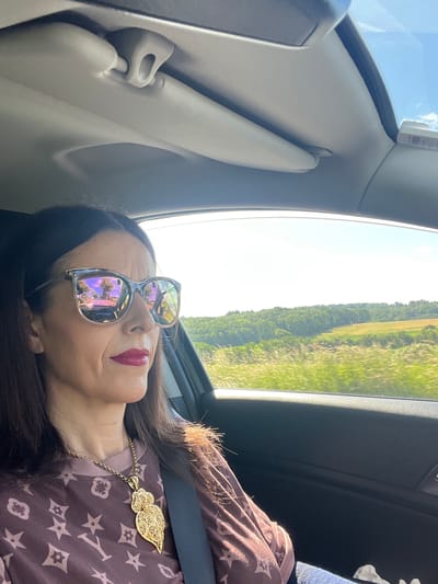A woman with dark hair, wearing sunglasses and a maroon t-shirt with a repeating star and flower pattern, is pictured from the chest up, inside a car. The sunglasses have a reflective surface showing a road and trees. She is wearing a large, intricate gold pendant necklace. A seatbelt is visible across her chest. The background shows a bright, sunny landscape with green trees and fields, suggesting an outdoor setting. The car's interior is visible, with a sun visor at the top of the frame. The image appears to be taken from the passenger seat. The scene is not specific to the city of Berg, Luxembourg; it depicts a general roadside view.
