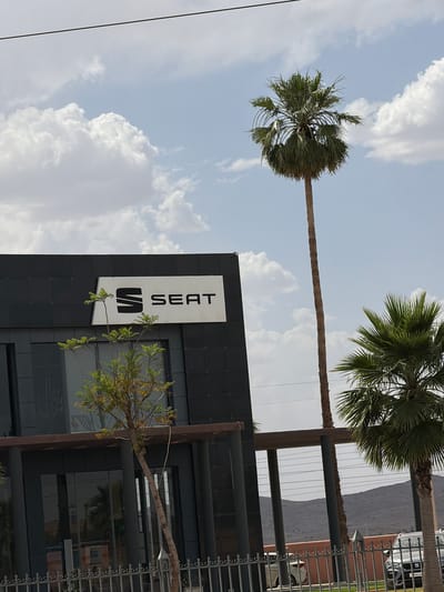 The image shows a building with a "SEAT" sign in Ouahat Sidi Brahim, Morocco. Two palm trees are visible. The sky is partly cloudy. A fence runs along the side of the building. Cars are parked nearby.