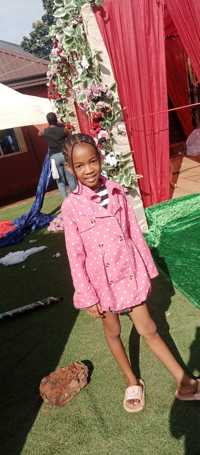 The image depicts an outdoor scene in Udi, Nigeria, featuring artificial green turf under direct sunlight. A young girl stands prominently in the foreground, angled slightly to the left, facing the camera with a smile. She has dark skin, braided hair, and wears a pink long-sleeved jacket with white polka dots over a striped shirt, blue shorts, and light-colored slide sandals. Her left knee is bent, and her right leg is extended.
In the mid-ground, an adult, viewed from behind, is bent over and manipulating piles of blue and red fabric on the turf. This individual has dark skin, braided hair, and wears a black top and blue jeans, with a white cloth tucked into the back of the waistband. Additional crumpled white fabric is on the ground nearby.
To the right, a vertical decorative structure is draped with red fabric and adorned with artificial flowers in white, pink, and red, intertwined with green leaves. More red drapes are visible behind this structure. A reddish-brown, irregularly shaped object rests on the turf near the girl's left foot, and a banded cylindrical object lies on the turf to the left. In the background, a building with a reddish-brown roof and a white canopy tent are visible, along with green trees against a clear sky. Strong shadows are cast on the turf by the subjects and structures.