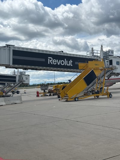 The image shows an airport tarmac at Vienna International Airport (VIE) in Schwechat, Austria, under a cloudy sky. A jet bridge with the "Revolut" logo is prominently displayed, connecting to an unseen terminal. To the right of the jet bridge, a yellow airstair with solar panels is positioned. The tarmac is marked with concrete barriers and painted lines. In the background, a plane with "LAUDA" written on its side is visible. A red fire extinguisher is placed on the tarmac near some airport equipment. The scene is static, with no visible people actively engaged in tasks.