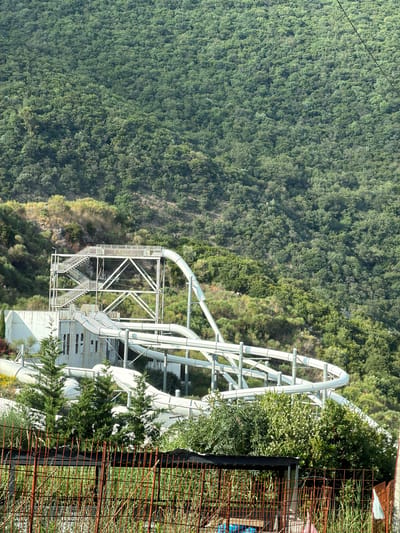 The image depicts an outdoor scene, possibly a water park or amusement park, built on a hillside overlooking a lush, green forest. A prominent white structure, which appears to be a roller coaster or a large water slide, dominates the right side of the frame. It features winding tracks and a complex metal support system. The structure is partially overgrown with vegetation, suggesting it might be abandoned or under construction.
To the right of the white structure, there is a rusty metal fence with a dense growth of tall grass and plants behind it. Some of the greenery around the structure is also quite dense, with trees and bushes growing up around it. The background is a vast expanse of dense, dark green forest that covers the hillside.
The lighting suggests it is daytime, with bright sunlight casting shadows. The weather appears to be clear. The overall impression is one of a slightly neglected or abandoned recreational facility set against a wild, natural backdrop. There are no people visible in the image. No text is legible. The location context provided is Seoce, Montenegro.