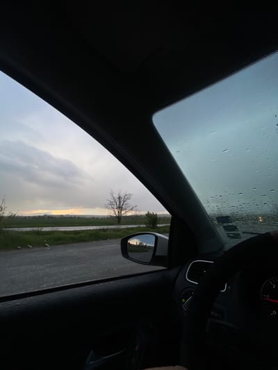The image shows a view from inside a car, likely the driver's perspective. The windshield has water droplets, suggesting recent or current rain. The steering wheel and part of the dashboard are visible in the upper right. The side mirror shows a reflection of a rural landscape with a bare tree, fields, and a road. The sky is overcast with grey and white clouds. The exact location in Prilep, Bulgaria cannot be determined based on the image alone. There is no presence of people in the picture.