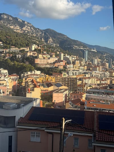 An elevated, wide-angle view captures a densely built urban landscape, identified as Beausoleil, France, looking eastward towards Monaco, under a bright blue sky with scattered white clouds. The immediate foreground features a terracotta-tiled roof with a prominent array of black solar panels. To its left, a flat, light-colored roof with a rectangular ventilation unit is visible. A wooden or concrete utility pole stands on the right, adorned with multiple black electrical cables and a downward-facing street lamp. Below the pole, a peach-colored building facade with a white-shuttered window is partially visible.
The mid-ground reveals a sprawling cityscape built into a steep hillside. Numerous multi-story residential and commercial buildings, varying in color from yellow, orange, and pink to beige and white, feature predominantly terracotta-tiled roofs. Several active construction sites are evident, marked by visible cranes. A distinctive, tall, slender skyscraper (likely the Tour Odéon) rises prominently in the mid-ground, right of center. Scattered green spaces and park areas are interspersed between structures.
The background is dominated by rugged, tree-covered mountains, with exposed rocky faces, ascending steeply under the clear sky. The urban development extends partly up these mountain slopes. No individuals or specific actions are discernible within the frame. The scene is illuminated by strong sunlight, casting distinct shadows, indicating either morning or late afternoon.