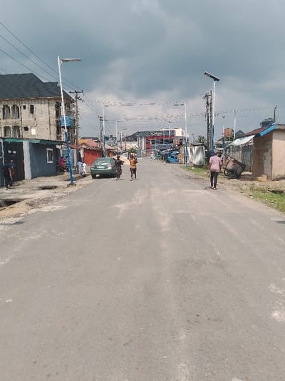 An asphalt road in Mgbuoshimini, Nigeria, extends into the distance under a heavily overcast sky. The road surface exhibits wear and minor repairs.

On the left, an open concrete gutter runs alongside the road. A small, blue-grey building with a dark roof stands near an unfinished, multi-story concrete building featuring exposed rebar and a dark gabled roof. Several individuals are clustered near these structures. A light green sedan is parked facing away from the viewer on the left side of the road. Utility poles with entangled wiring and white pole-mounted streetlights with solar panels are prominent features on both sides of the street.

In the middle of the road, two individuals walk towards the viewer. The leading person carries a large, light-colored object on their head. A second person, slightly behind and to their right, also carries an item on their head.

On the right side of the road, a man in a light pink shirt and dark trousers walks away from the viewer. A small, tan building with a blue roof is visible, along with an overturned, dark-colored wheelbarrow-like object on the dirt shoulder. Further down, another small, tan building with a flat roof is present.

In the background, the street is lined with additional buildings, including a multi-story structure with red and blue accents on the distant right. Numerous streetlights continue along the road's length.