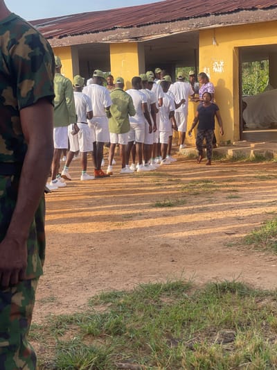 The image depicts an outdoor scene at what appears to be a facility in Emure-Ekiti, Nigeria. In the foreground, the left side of the frame is occupied by the back of an individual wearing a camouflage military uniform.

The main subject of the image is a line of young men in white shirts and shorts, some wearing green caps, standing in a dusty area. They are facing towards a yellow building with a corrugated iron roof. Some of the individuals are looking towards the building.

Near the entrance of the yellow building, two women are present. One woman, wearing a purple and white patterned top, is interacting with the individuals in the white uniforms. Another woman, in a dark top and plaid pants, stands with her arms slightly out.

The environment is characterized by a dirt ground with patches of grass, and the yellow building appears somewhat aged. Sunlight casts shadows across the ground, indicating it is daytime. The overall scene suggests an organized gathering or activity involving the individuals in uniform and the women.