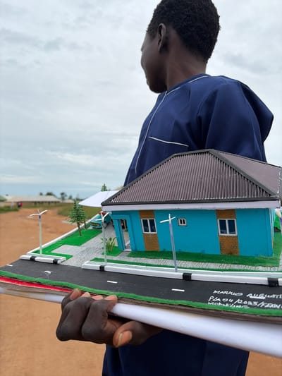A person wearing a blue kaftan stands outdoors in Wumat Central, Nigeria, holding a diorama of a blue house with a brown, corrugated roof. The diorama features a small driveway, green grass, miniature trees, and streetlights. The house has brick-patterned accents around the windows and doors. The person's right hand is visible, holding the edge of the diorama. In the background, there are buildings and trees under a cloudy sky. There is text written on the edge of the diorama that appears to read "MARKUS AUGUSTIN, PLO 4/2023/FEXIVO MODELLING".
