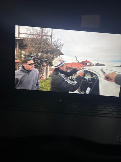 A medium-distance shot depicts two individuals outdoors in what appears to be a residential or semi-rural setting. One man, wearing a grey hoodie with a Nike logo and sunglasses, stands in the foreground. Behind him, another man, wearing a black patterned jacket and a white hat, is seen interacting with a white vehicle. A hand, possibly belonging to a third person, is holding an object near the top of the car door. In the background are buildings, a fence, and trees. The photo was taken in Kalchevo, Bulgaria.