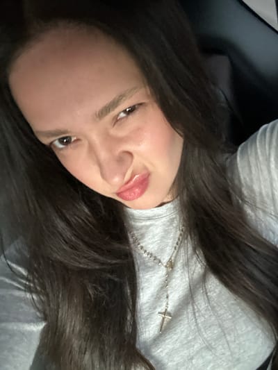 A close-up image features a single individual, likely female, with long, dark brown hair and a light complexion. The subject is positioned facing the camera, with lips pursed in a puckered expression and a slight furrowing of the brows. She wears a light grey, long-sleeved garment and a silver-colored rosary necklace with a crucifix pendant. The background is dark and indistinct, with a dark, possibly upholstered surface visible behind her right shoulder. The specific location within San Bernardino, United States, is not discernible from the provided image details.