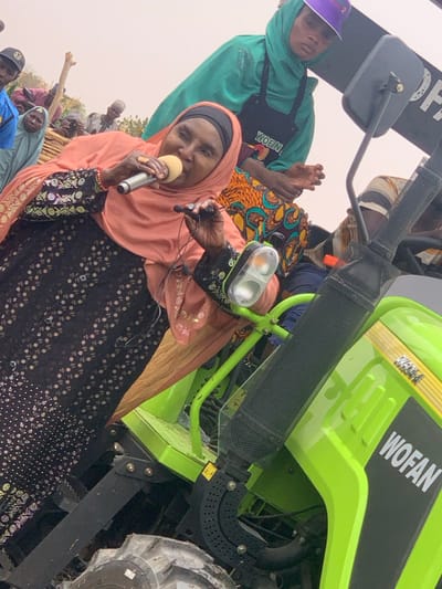 A woman wearing a peach-colored headscarf and a black, sequined dress is speaking into a microphone while seated on a bright green tractor in Ringimawa, Nigeria. The tractor, a WOFAN model, has a black exhaust pipe and a black rearview mirror. The woman is smiling and appears to be addressing a crowd, but the crowd is not visible in the image. To the woman’s left, another woman in a teal headscarf is seated on the tractor with her hand on the woman's back. The woman in teal is wearing a black apron with the word "WOEIN" printed on it. The woman in teal is looking at the microphone. The scene is outdoors, with a hazy sky in the background. The photo was taken in Ringimawa, Nigeria, which is located at approximately 11.666° N, 8.415° E.