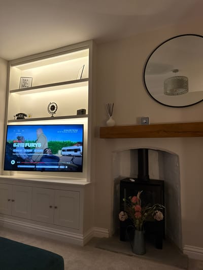 The image displays a cozy, well-decorated living space, likely a living room, illuminated by warm indoor lighting, suggesting evening or nighttime. The scene is dominated by a white built-in entertainment unit and shelving system, and a fireplace niche.

On the left, the built-in unit features open shelves with integrated LED strip lighting, showcasing various decorative objects. These include a vintage-style clock, an old-fashioned rotary phone, a small white daisy ornament, and a framed piece of text reading "Mek sen home". A television is centrally positioned within this unit, displaying content from Netflix. Below the shelves and TV, white cabinets with recessed panel doors provide storage.

Above the built-in unit, a rustic wooden beam is mounted horizontally on the wall. To its left, a large round mirror with a black frame reflects a ceiling light fixture with an ornate, metallic lampshade. Below the beam, a white ceramic diffuser with black reeds rests on a small shelf.

To the right, a white-painted fireplace niche houses a modern black faux log burner. A light blue pitcher containing a bouquet of artificial flowers in shades of pink and white is placed on top of the burner. The niche features decorative molding along its edges. A fragment of a dark-colored sofa and light-colored carpet are visible in the foreground.

The television screen shows the Netflix series "At Home With The Furys", specifically "S2: E2 'I'm Gonna Tell You a Secret'". On-screen text includes "NETFLIX", "OPTIONS", feedback prompts like "Not for me", "I like this", "Love this!", and "Enjoying this? Rating helps us know we should recommend more like this". Audio and subtitle options like "English (United States) [Original] with Subtitles" are displayed, along with timestamps "20:12" and "17:13". The decor and comfortable setting suggest a modern British home, aligning with the Harrogate, United Kingdom context.