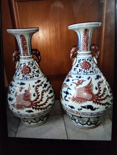 A pair of blue and white porcelain vases with red phoenix and floral motifs, each with two elephant-shaped handles, are displayed against a wooden paneled background. The vases are situated on a tiled surface. The image is framed by a dark border. The location of Kualaraja, Indonesia, is not depicted in the image.