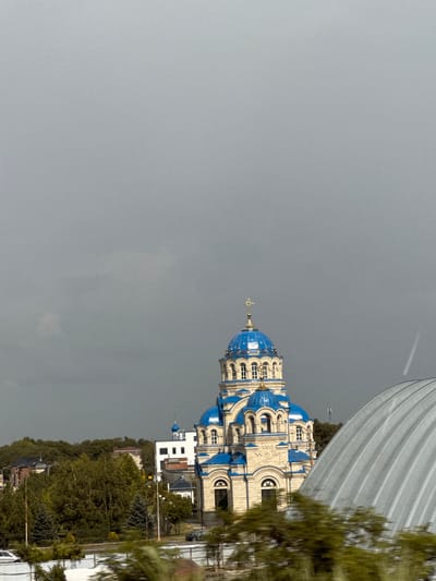 The image shows a building with blue domes, resembling a church, under a cloudy sky. The building is ornate, with light-colored walls and arched windows. Several blue domes are visible, with a prominent one at the top adorned with a cross. To the right of the building, part of a large, curved, white structure with horizontal lines is visible, potentially a roof or awning. A tree with green leaves is in the foreground on the right, partially obscuring the view of the building. The overall scene suggests an outdoor setting, possibly in a town or city, given the presence of other buildings and what appears to be a road or pathway in the distance. The sky is overcast and gray, indicating a cloudy day. The image appears to be taken from a slightly elevated perspective. There is no visible text in the image.