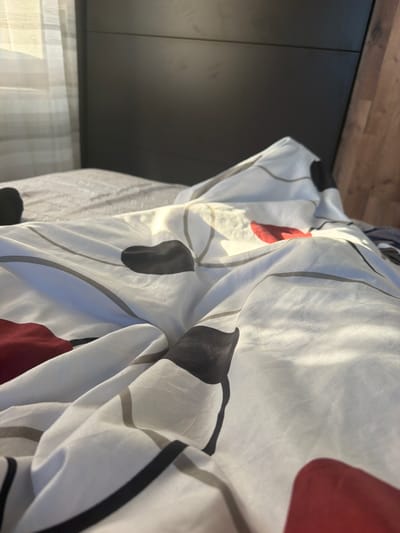 A close-up shot shows a bed with crumpled white sheets patterned with gray, black, and red organic shapes. To the left of the sheets, there are gray cabinets with a wooden top and a window with white curtains. The location of the image is unclear and does not relate to the city of Petarch, Bulgaria. There are no people, subjects, objects, actions, or interactions described in this image.