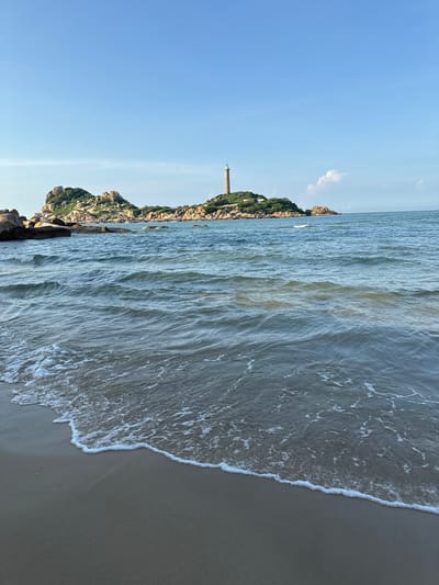 The image depicts a serene coastal landscape with a prominent lighthouse situated on a rocky island. The lighthouse, a tall, slender structure, stands atop a hill covered in lush green vegetation and scattered rocks. The island itself is a cluster of rugged, natural formations jutting out from the calm, blue ocean. In the foreground, gentle waves lap against a sandy beach, creating a soft white foam line. The sky above is a clear, vibrant blue, with a few wispy clouds adding a touch of softness to the horizon. The overall scene suggests a peaceful and picturesque day by the sea. While the specific location is identified as Thuận Thành, Vietnam, the image captures a natural, undeveloped coastal scene rather than a bustling urban environment. There are no people or visible signs of human activity beyond the lighthouse itself.