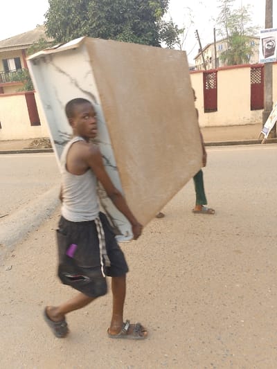 A young boy walks on a street carrying a large, heavy sheet of material, likely plywood or similar board. He wears a tank top, dark shorts, and sandals, and has a worried expression on his face. Another person, presumably helping, is partially visible behind the boy, also carrying the sheet. The scene appears to be on a residential street in Africa, with houses and buildings visible in the background.