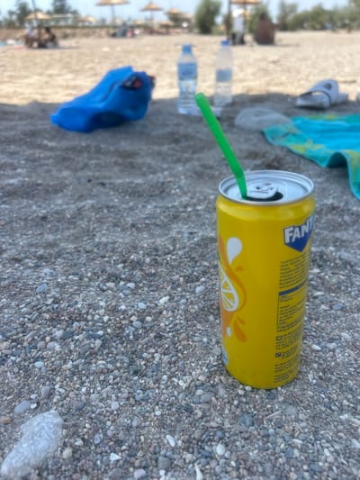 A bright yellow can of Fanta, with a green straw inserted into its opening, rests on a pebbly beach. The can is positioned on the right side of the frame, with the lower half of the can and the pebbles filling the foreground. The background is slightly blurred, showing a beach setting with umbrellas, people in the distance, and two plastic water bottles on the sand. A blue object, possibly a bag or towel, is visible to the left of the can. The scene appears to be a casual day at the beach.