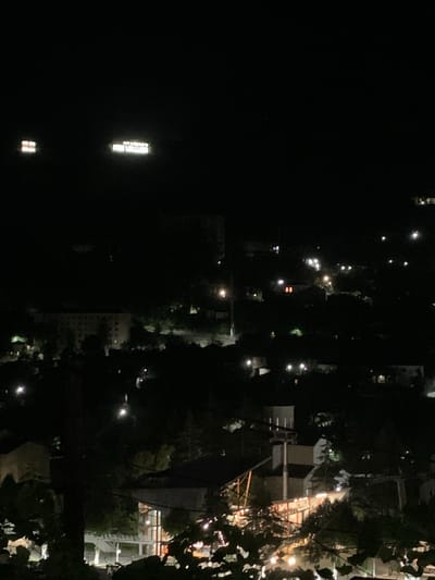 The image is a nighttime aerial view of the city of Chiatura, Georgia. The darkness of the night is punctuated by various lights, indicating the presence of buildings and infrastructure. In the upper left quadrant of the image, two illuminated structures are visible. One appears to be a building with lit windows forming a grid pattern. To its right, a larger, rectangular illuminated sign displays text in what appears to be a sans-serif font, with individual letters clearly distinguishable. The text on this sign reads "CHiatura" on the top line and "CITY CENTER" on the bottom line.

Further into the cityscape, numerous scattered lights indicate residential and commercial areas. Some lights appear as streetlights, illuminating roads or pathways, while others emanate from the windows of buildings, suggesting occupancy. The landscape is hilly, with trees and vegetation interspersed among the built-up areas, creating a texture of light and shadow. The overall impression is of a city at rest, with only the ambient lighting and the illuminated signs providing a sense of its presence in the darkness. The image is slightly blurry, consistent with a nighttime photograph taken without a tripod or with a low-light setting.