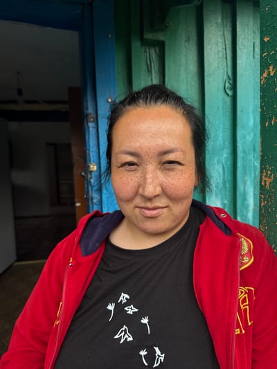 A medium shot, taken outdoors, shows a woman with dark hair and freckles smiling at the camera. She is wearing a red hooded jacket over a black t-shirt with a white bird graphic. The background consists of a wooden structure painted in teal and blue. A doorway with dark interior is visible to the left. The image captures the woman from the chest up, with the focus on her face and attire. The setting appears to be rustic, possibly a residential dwelling in Yabogan, Russia, given the painted wooden structures commonly found in such locations.