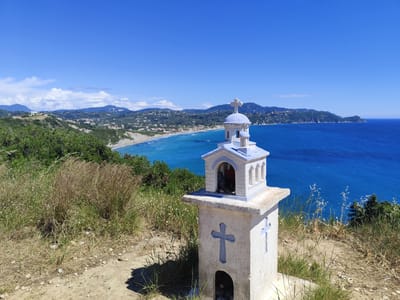 This image presents a stunning panoramic view of a coastal landscape, likely in Agios Stefanos, Greece, from an elevated vantage point.
In the foreground, a small, whitewashed miniature chapel or shrine stands prominently on a dirt path. It features a light blue domed roof crowned with a cross, and two smaller side turrets also topped with crosses. The main structure has two light blue crosses embossed on its sides and an arched opening at its base, with another larger arched opening on its front revealing a dimly lit interior. The immediate ground is covered in dry dirt and various grasses, both green and dried.
Beyond the shrine, the midground reveals a lush, green hillside sloping down to a long, sandy beach. The coastline is dotted with numerous buildings and settlements, indicative of a vibrant coastal village. The sea closest to the shore exhibits a vibrant turquoise color, gradually deepening to various shades of blue further out.
The background features the expansive, deep blue waters of the sea stretching to the horizon. Along the coastline, undulating green mountains and headlands extend into the distance. The sky above is a brilliant, clear blue, with only a few faint white clouds visible in the far distance over the mountains on the left.
The scene is bathed in bright, clear daylight, suggesting a sunny day, possibly midday or early afternoon, given the short, distinct shadows cast by the shrine. The overall impression is one of serene natural beauty, typical of the Mediterranean region. There are no people visible, and no discernible text appears in the image.