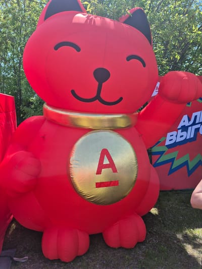 An inflatable red cat figure, styled as a Maneki-neko, is depicted outdoors in the city of Kansk, Russia. The large, red inflatable features black crescent-shaped eyes, a black triangular nose, and a black smiling mouth. Its inner ears are also black. A golden inflatable collar encircles its neck, and a prominent golden inflatable medallion with a red capital letter "A" and a horizontal red line beneath it is centered on its belly. The cat's right paw is raised, consistent with the traditional Maneki-neko gesture. Its lower body and three-lobed feet are visible, resting on green grass. In the background, lush green tree foliage is visible under a clear sky. To the right of the red cat, a section of another large, red inflatable object is partially visible. This background inflatable displays white Cyrillic text, reading "АЛЫ" and "ВЫГ", likely fragments of longer words, above zigzag patterns in green, blue, and black. A small portion of a human limb is visible on the extreme right, and a grey fabric anchor is on the grass at the bottom left.