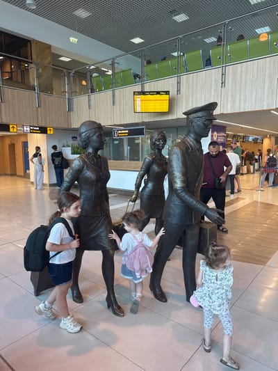 In the city of Bubuieci, Moldova, a group of people are observed in an airport terminal. In the foreground, two young girls, one with a black backpack and the other with a pink backpack, are interacting with bronze statues depicting a man and a woman in formal attire, possibly representing airport personnel or historical figures. The statues are positioned next to luggage. Behind them, other travelers can be seen, some with luggage, and a departure board displaying flight information. The terminal features modern architecture with a metallic ceiling, large windows, and seating areas in the background. Signage for restrooms and an information desk is visible.