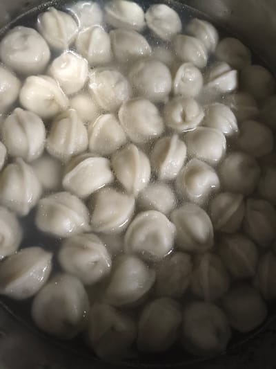 The image displays a close-up overhead view of a pot filled with numerous dumplings submerged in simmering water. The dumplings are uniformly pale and have a rounded, slightly pleated shape, typical of boiled dumplings. Small bubbles are visible on the surface of the water, indicating the cooking process. The lighting is diffused, highlighting the texture of the dumplings and the clarity of the water. The edges of a dark-colored pot are visible at the bottom and left of the frame. There are no people, animals, or outdoor elements present in the image. The context of Galonki, Poland, is not visually represented and cannot be inferred from the provided image.