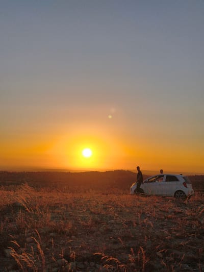 This vertical, eye-level shot captures a breathtaking sunset scene in Be'er-Sheva, Israel. The sky transitions from a soft blue at the top to a vibrant orange and yellow as it nears the horizon, where a brilliant sun is prominently displayed. A hazy glow surrounds the sun, creating a surreal and warm atmosphere.
In the mid-ground, a small white car is parked on a rocky, arid hillside covered with dry, golden grass that is illuminated by the setting sun. Two people are visible near the car. One, silhouetted in black, stands to the left of the vehicle, leaning against its side. Another person, less clearly defined, is seated inside the car. The landscape beyond the immediate foreground is a rolling expanse of hills and sparse vegetation, suggesting a semi-arid or desert environment characteristic of Be'er-Sheva.
The warm, golden light casts long shadows and highlights the textures of the dry grass, creating a sense of natural beauty and tranquility. The scene evokes a feeling of peacefulness and a connection with nature, likely capturing a moment of pause and appreciation for the end of the day. The time of day is clearly sunset, with the warm, low-angled light. No visible text is present in the image.